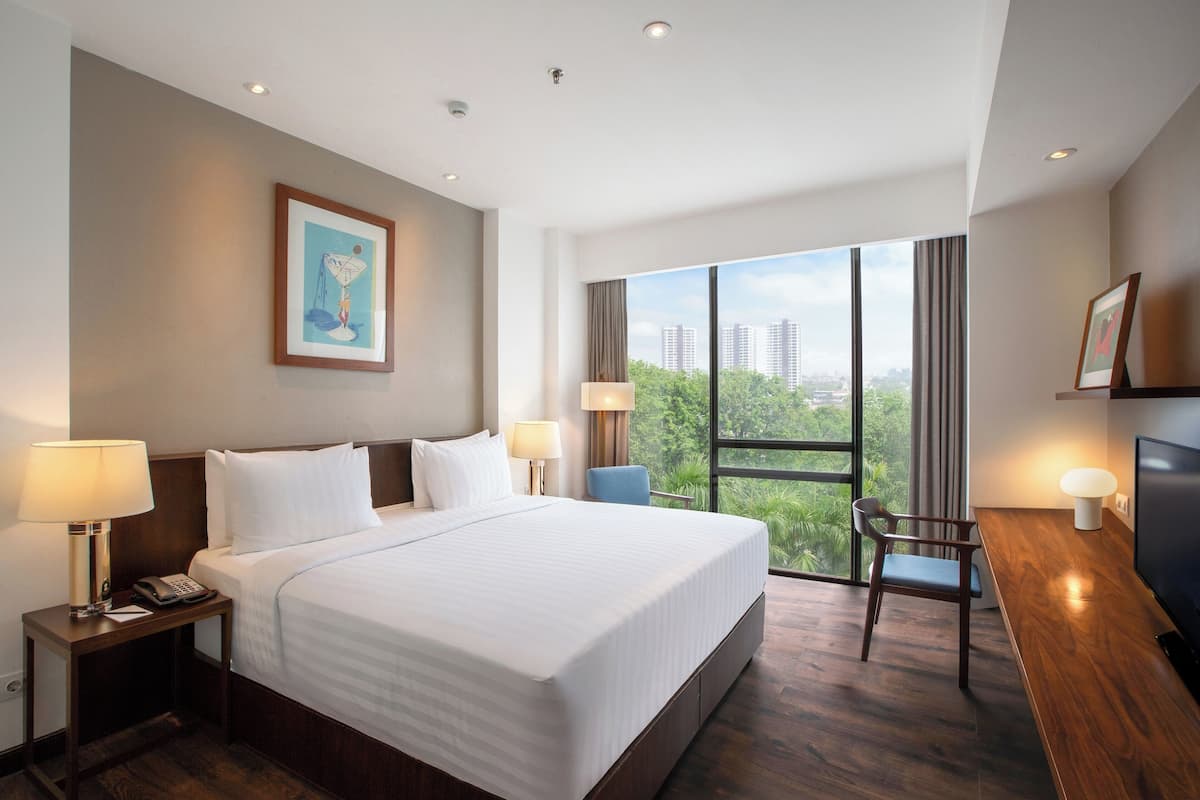A hotel room with a large bed, two bedside tables with lamps, a desk with a chair, and a large window offering a city view.