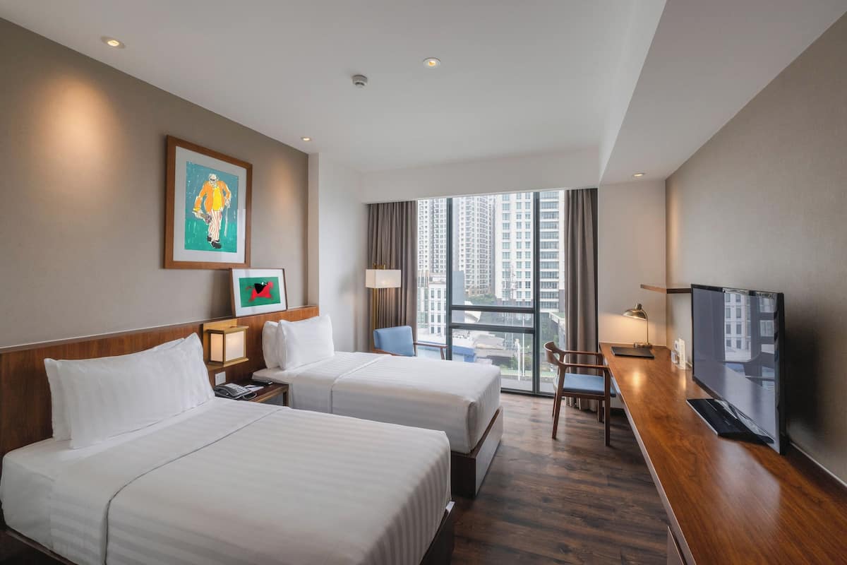 A hotel room with two beds, a desk, a TV, and a large window with a city view.