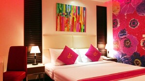 Une chambre d’hôtel avec un lit, deux oreillers ornés d’un logo, une table de chevet avec une lampe, une chaise et un tableau abstrait aux couleurs vives accroché au mur.