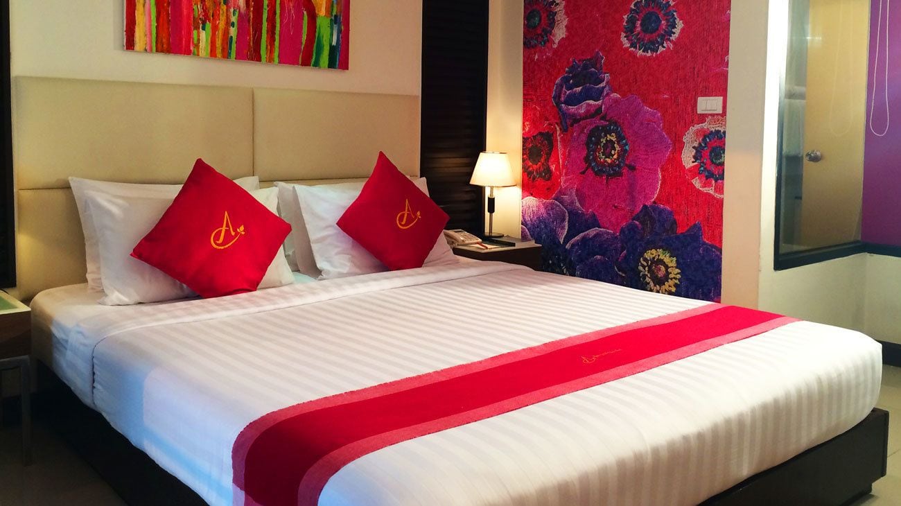 A hotel room with a large bed, two red pillows with a logo, a nightstand with a lamp, and a colorful wall tapestry.