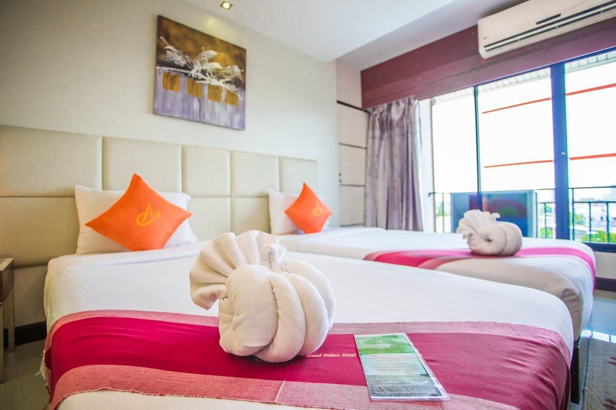 Ein Hotelzimmer mit zwei Betten, jedes mit einer roten Tagesdecke, weißen Kissen und einem dekorativen Handtuch. An der Wand hängt ein Gemälde und über dem Fenster befindet sich eine Klimaanlage.