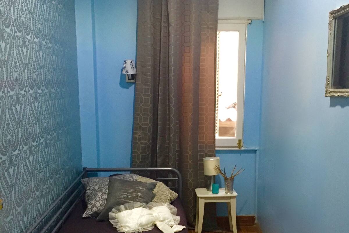 Quarto pequeno e compacto com uma cama de solteiro, mesa de cabeceira, janela com vista e paredes com padrão.