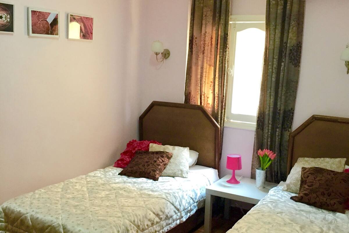 Quarto com duas camas de solteiro, uma pequena mesa de cabeceira com abajur rosa e uma janela com cortinas.