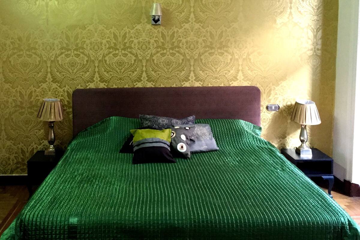Uma cama com colcha verde, duas mesas de cabeceira com abajures e um papel de parede estampado.