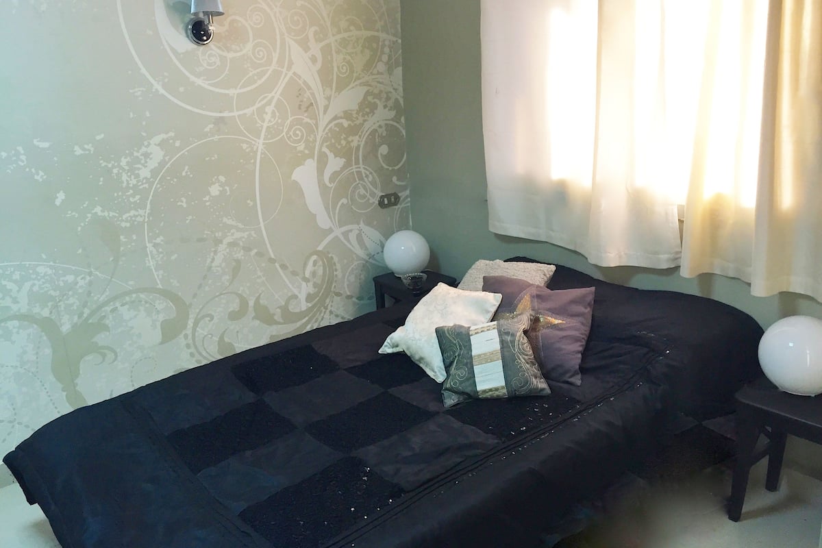 Quarto com cama, almofadas, mesa de cabeceira, abajur e parede com padrão decorativo.