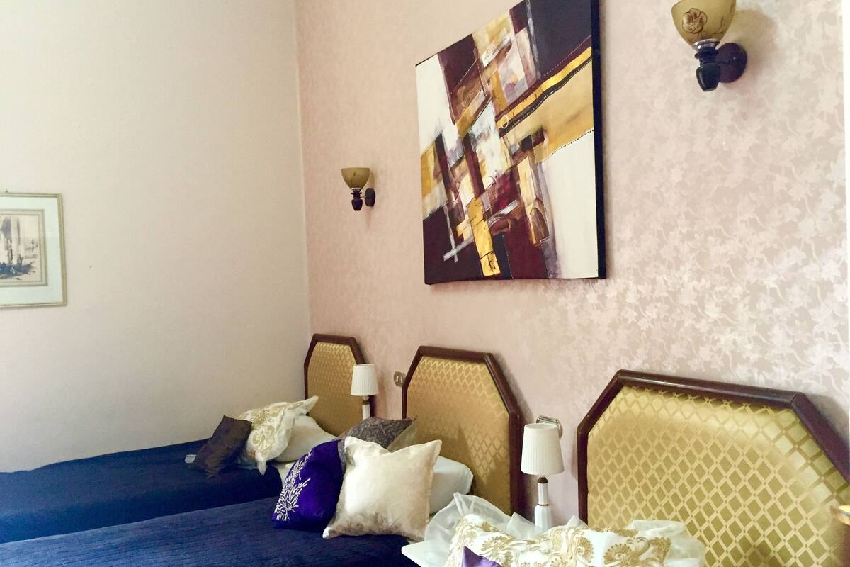 Quarto de hotel com duas camas, cada uma com cabeceira, luminária na parede e um quadro acima das camas.