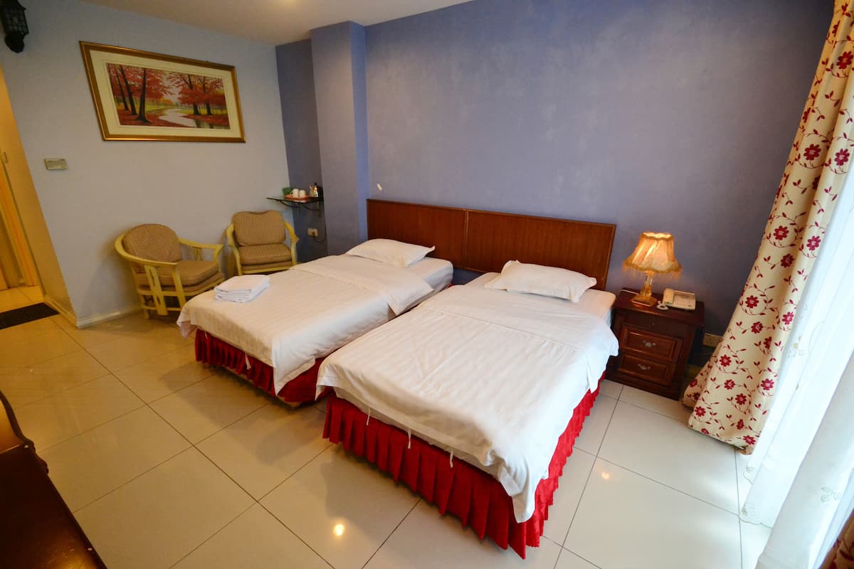 Une chambre d’hôtel avec deux lits, une tête de lit en bois, une table de chevet avec une lampe, une chaise et un tableau au mur.