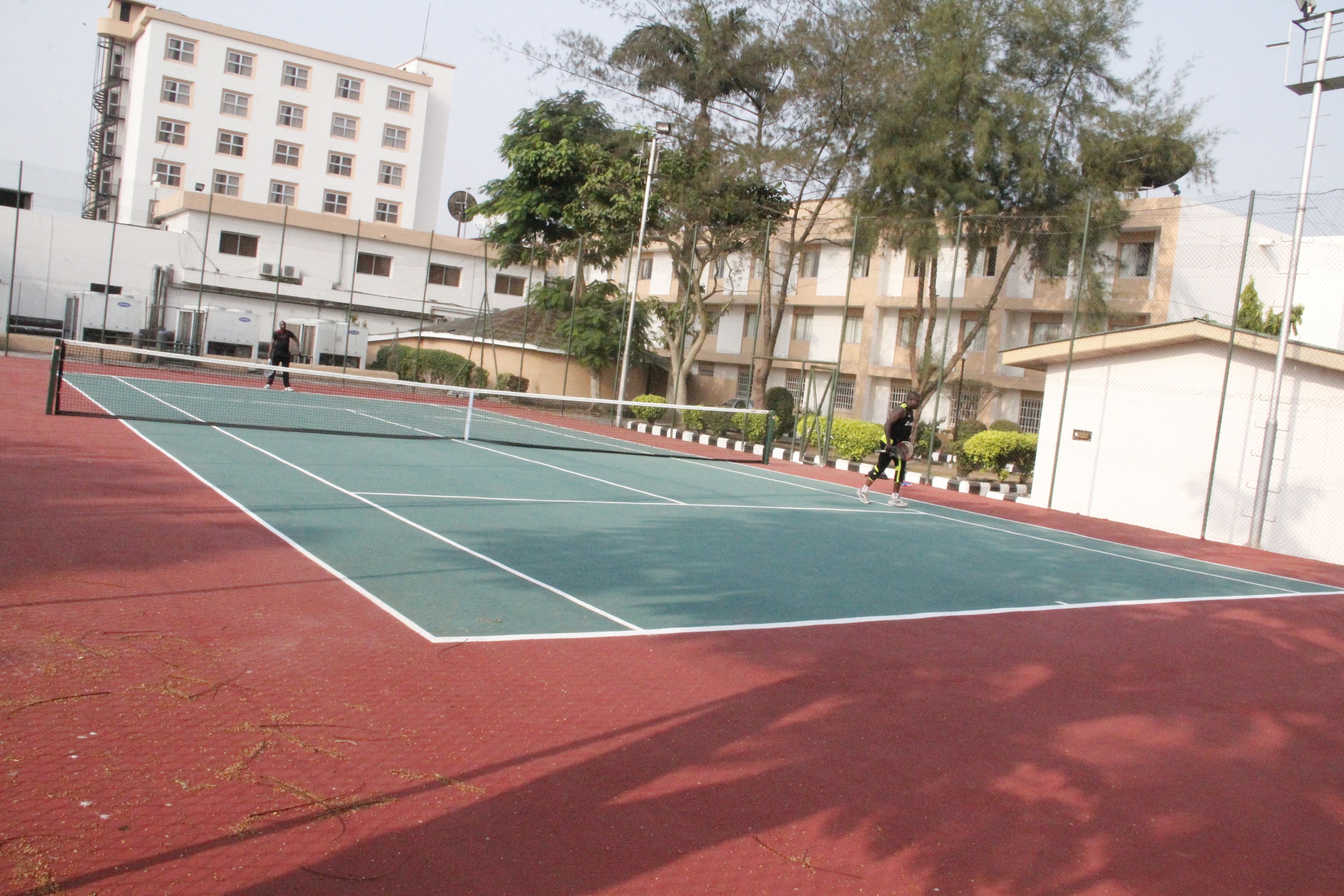 An outdoor tennis court with a net, surrounded by trees and a building in the background.
