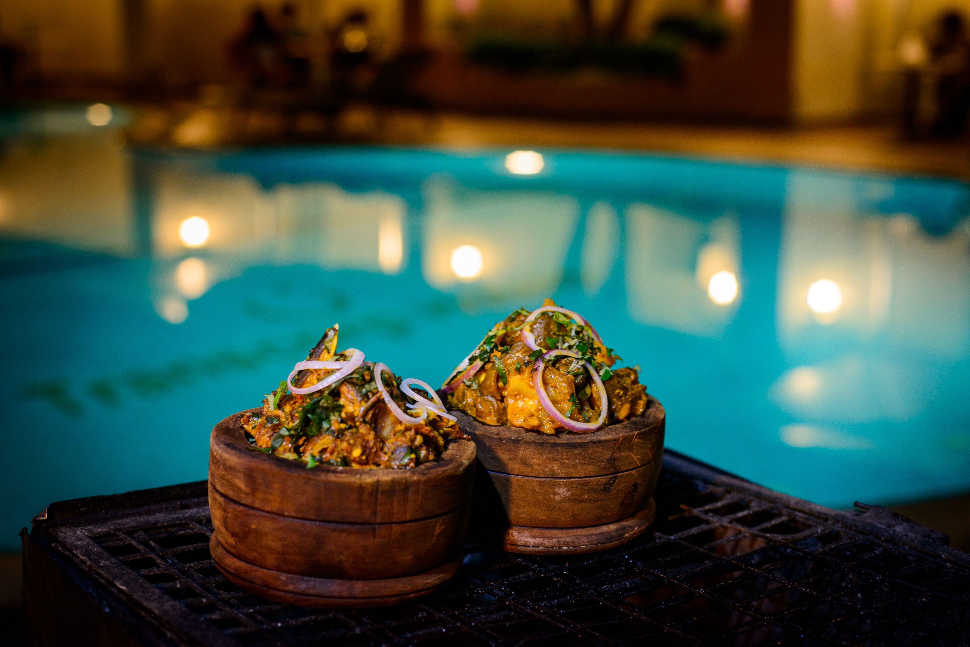Two wooden bowls of food on a grill with a pool in the background.
