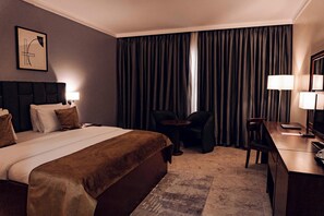 A hotel room with a large bed, a desk, two chairs, a small table, a lamp, and a window with curtains.