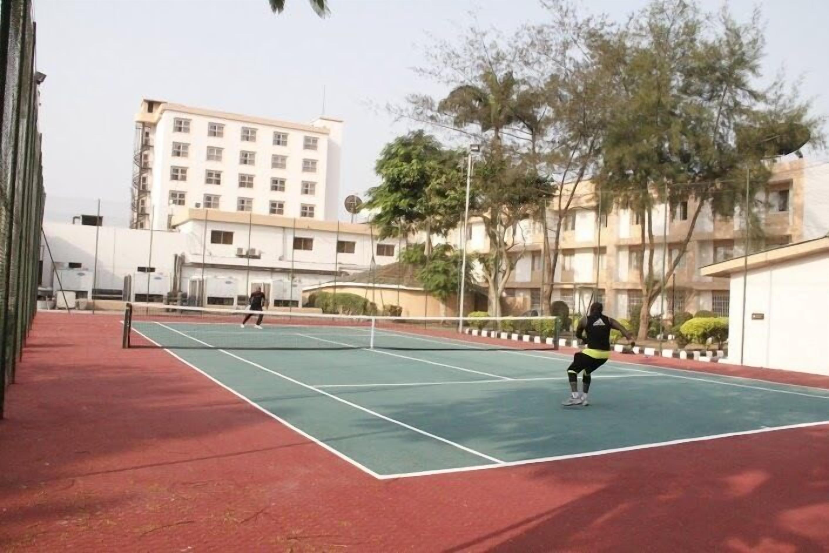 Una cancha de tenis al aire libre con dos jugadores, rodeada de edificios y árboles.
