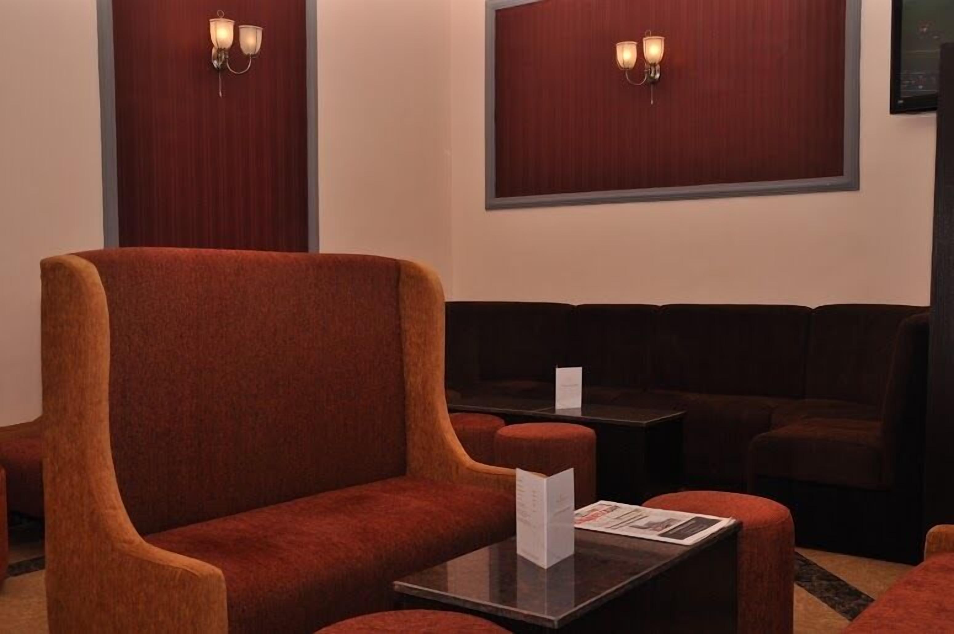 Un área de estar con sillones y sofás tapizados en rojo, una pequeña mesa con un menú, y lámparas fijadas en la pared.