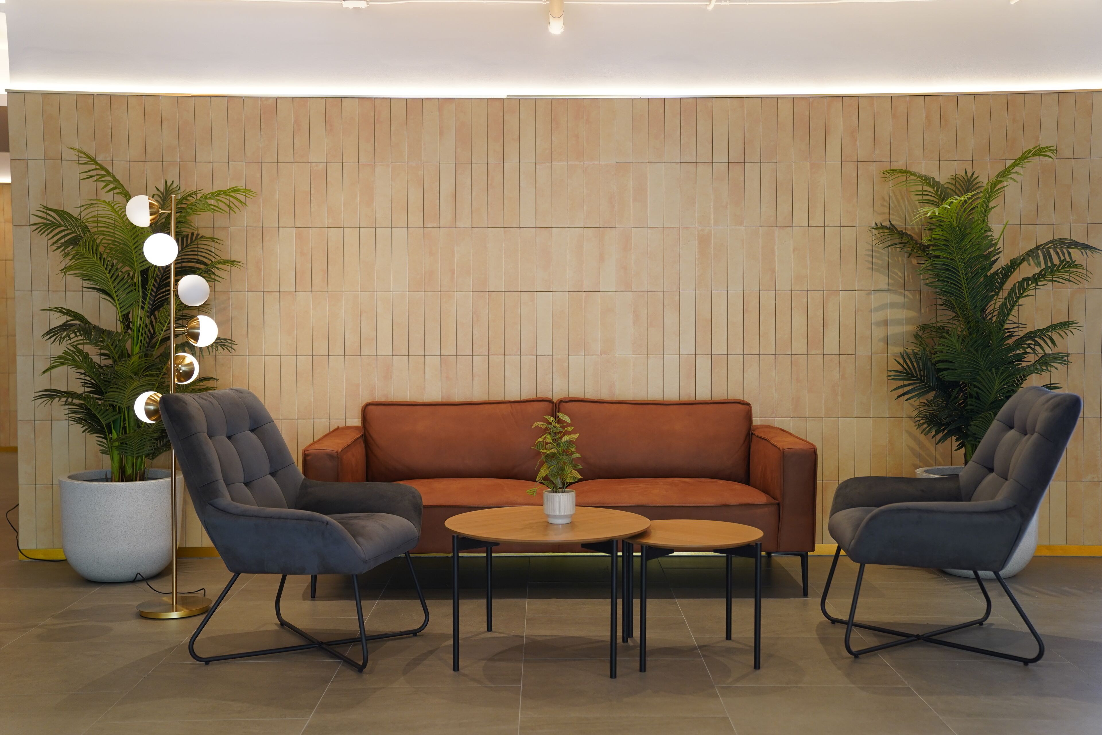 A modern lounge area with a brown sofa, two armchairs, and a coffee table. There are two potted plants and a floor lamp with multiple lights.