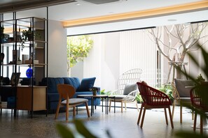Un salon moderne avec un canapé bleu, une table basse en bois et un fauteuil rouge. On y trouve une bibliothèque remplie de divers objets et une grande fenêtre qui laisse entrer la lumière naturelle.