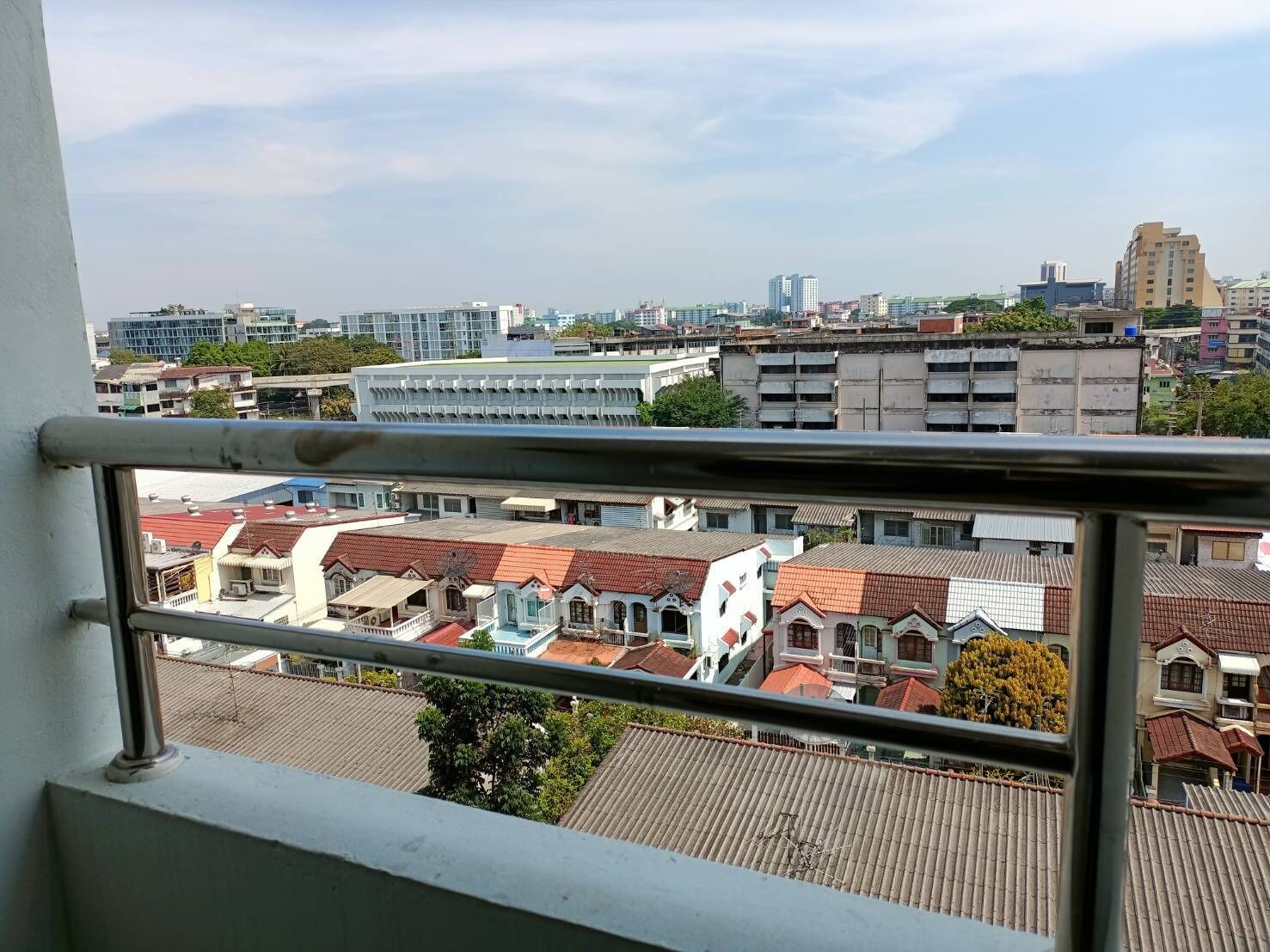 A view from a balcony with a metal railing, overlooking a cityscape with a mix of modern and traditional buildings, including a prominent white building with a red roof.