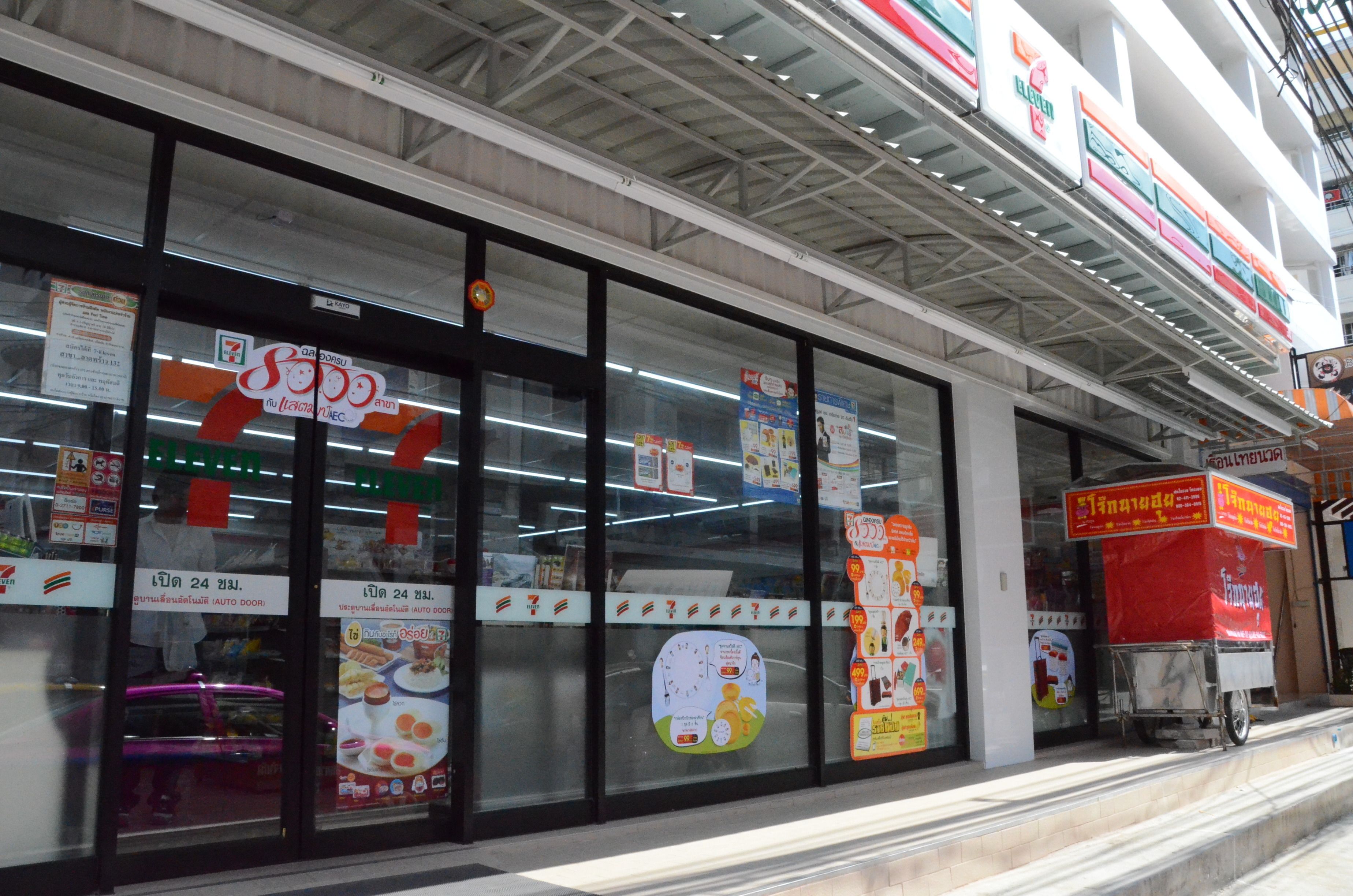 Suuri 7-Eleven-myymälä, jossa on suuret lasi-ikkunat, erilaisia mainoskylttejä ja punainen jakelukärry sen ulkopuolella.