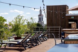 Una terraza en la azotea con sillones de sol, plantas en macetas y guirnaldas luminosas.