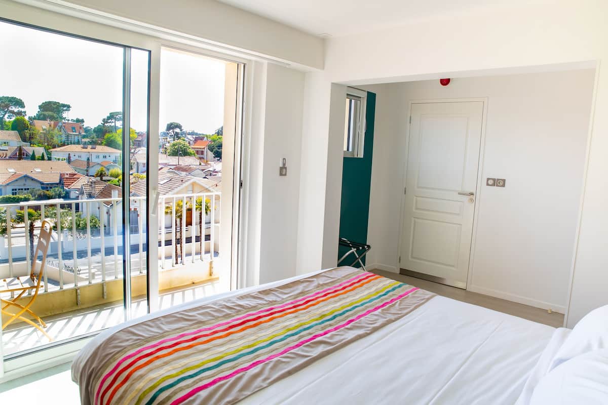 A bedroom with a large bed, a balcony with a chair, and a view of houses.
