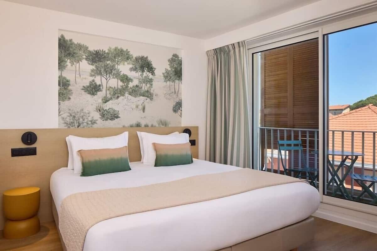 A hotel room with a bed, a painting on the wall, a balcony with chairs, and a view of buildings.