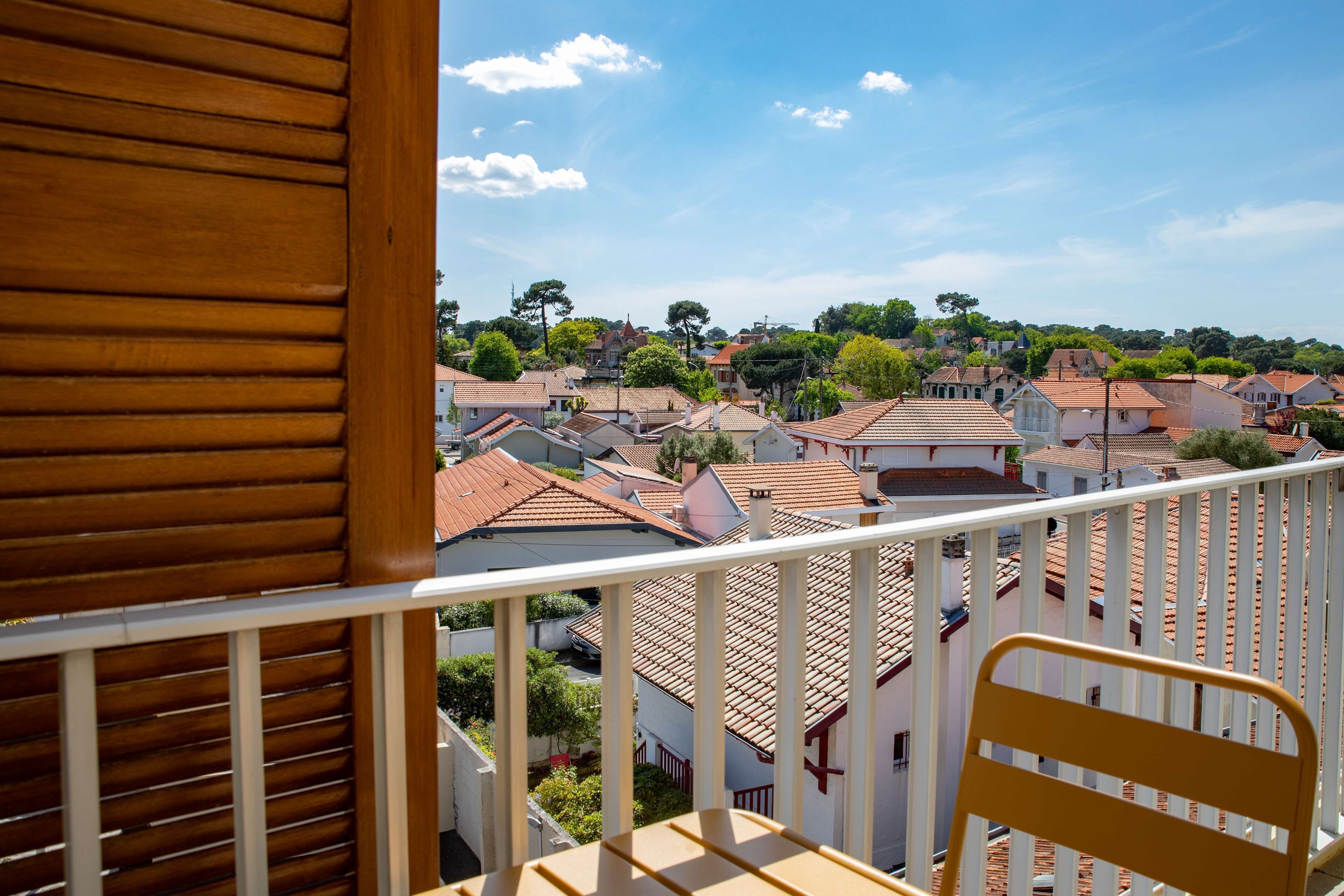 Un balcón con barandilla de madera y una silla amarilla, con vista a un pueblo con tejados rojos y zonas verdes.