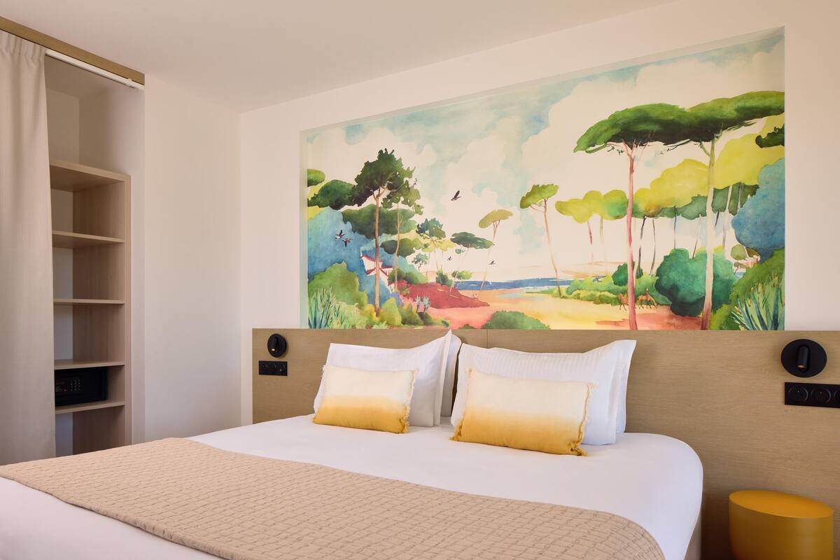 A hotel room with a bed, a large landscape painting on the wall, a wardrobe, and a bedside table.