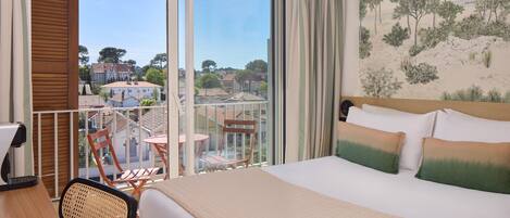 Une chambre d’hôtel avec un lit, un bureau, une chaise, un balcon donnant sur des immeubles et un tableau au mur.