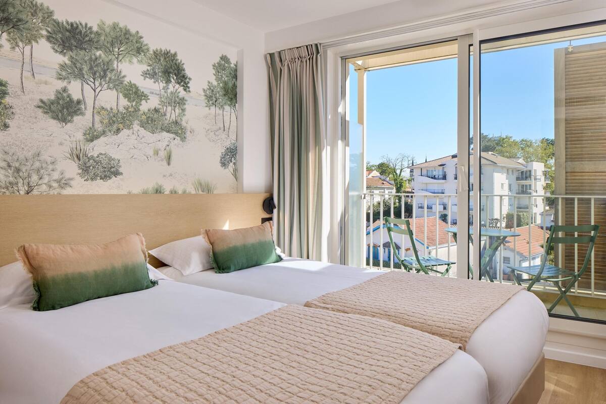 A hotel room with two beds, a balcony with a view of buildings, and a painting on the wall.