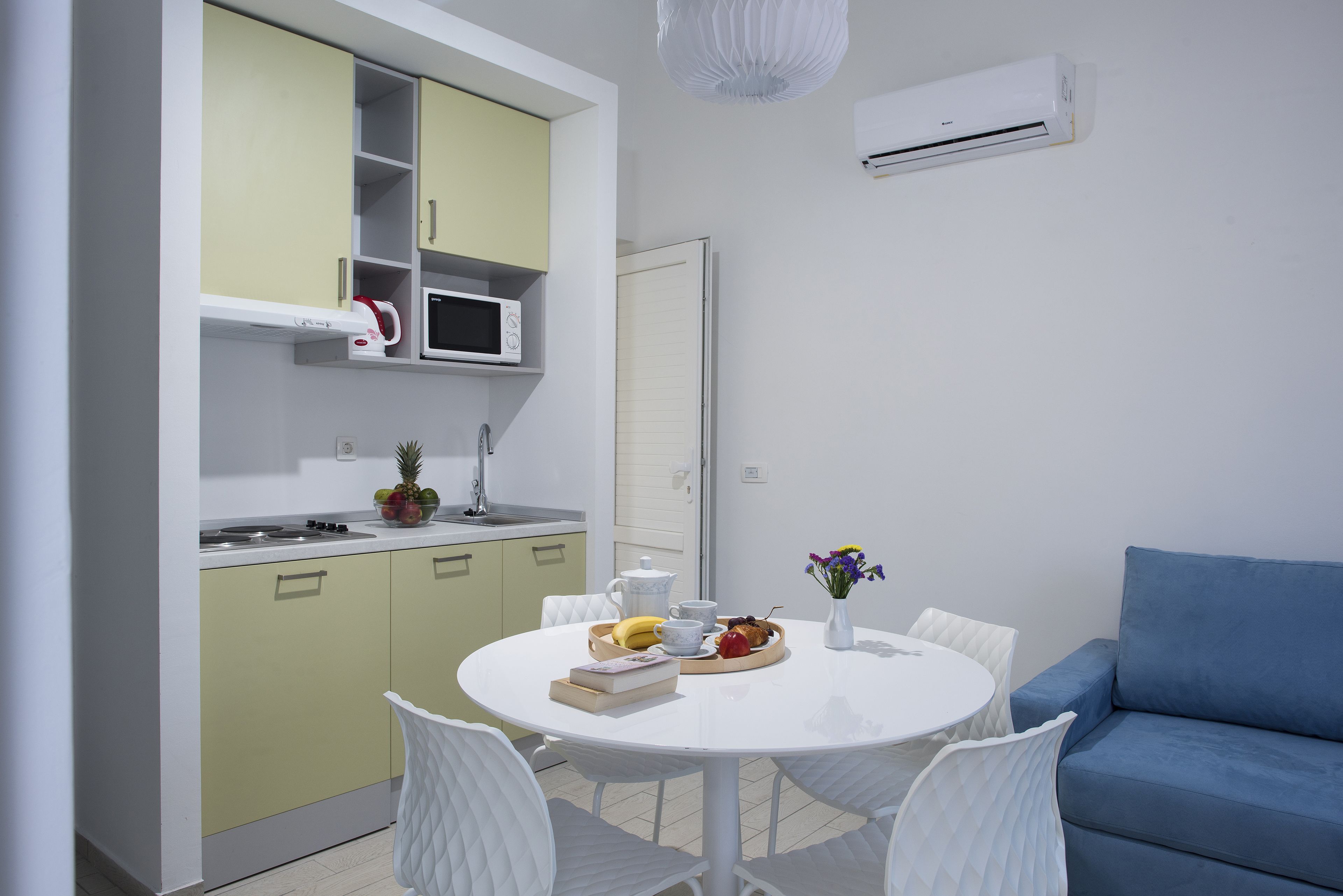 A compact kitchen with a microwave, a kettle, and a fruit bowl on the counter. There is a dining table with chairs and a blue sofa in the living area.