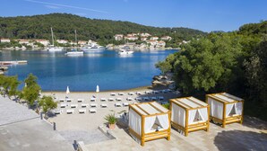 Un hotel frente al mar con habitaciones junto a la playa, un puerto deportivo y vistas a un pueblo en la colina.