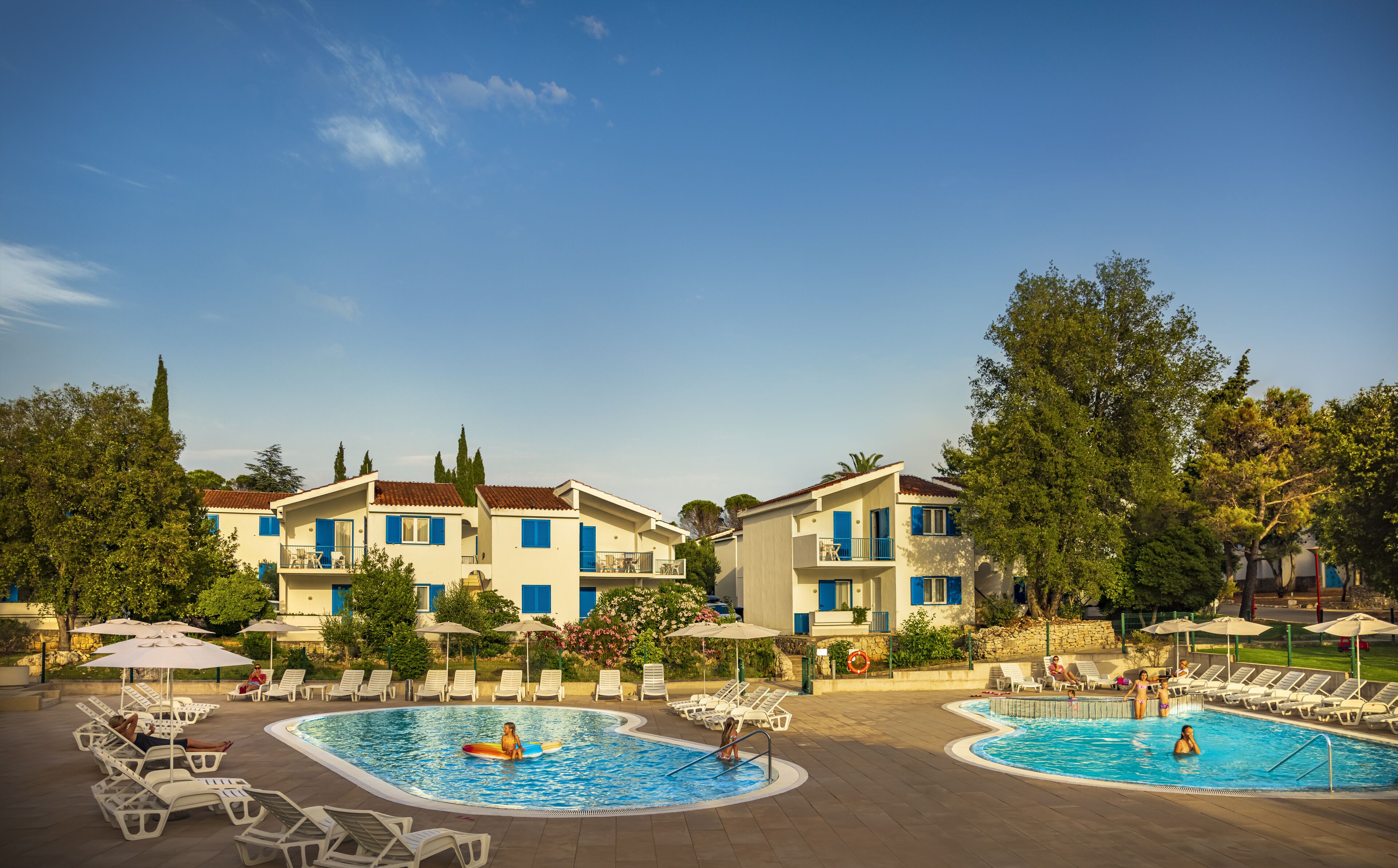 Ein Resort mit zwei Schwimmbecken, Liegestühlen und einem Gebäude mit blauen Fensterläden.