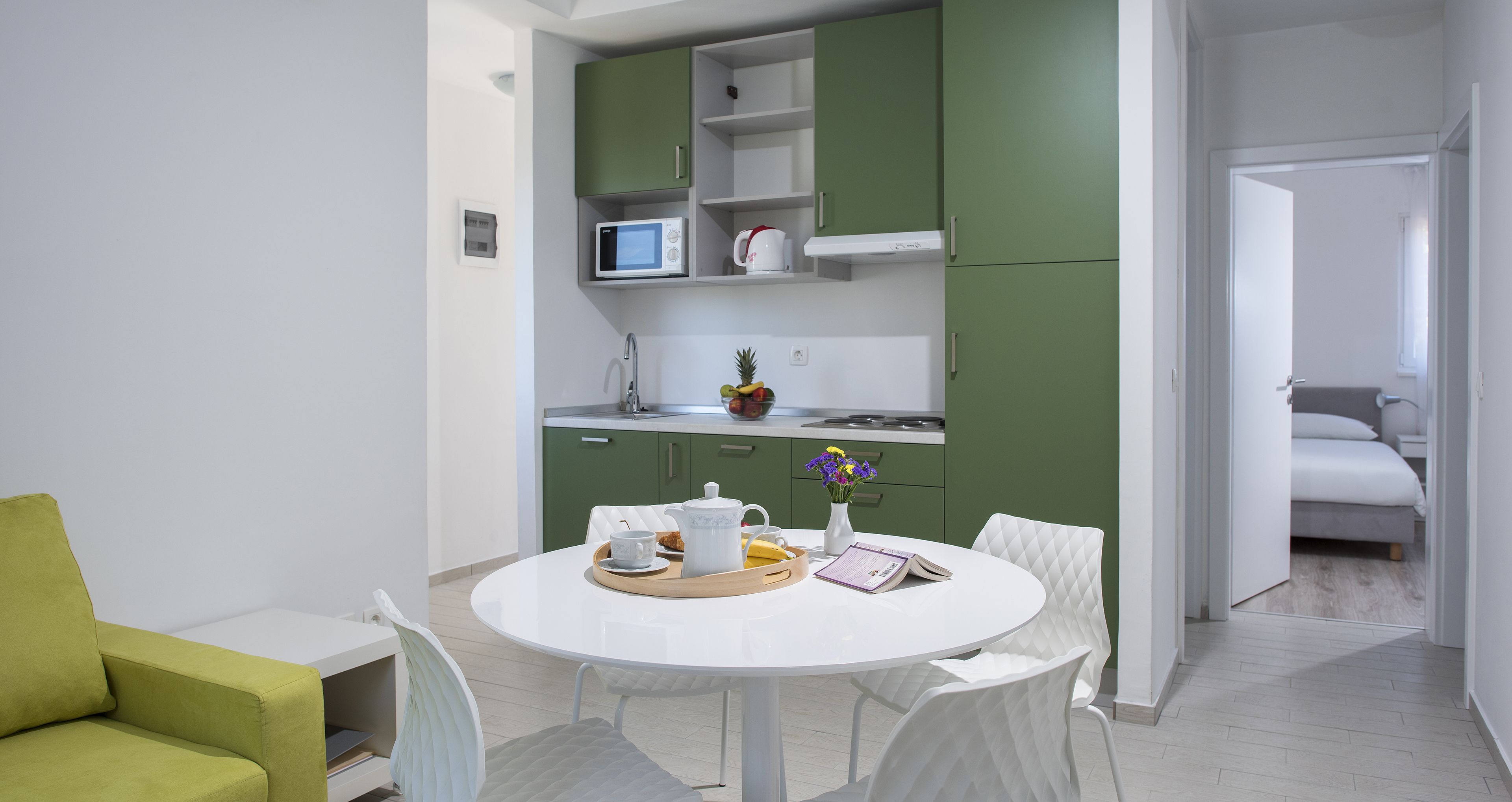 A modern kitchen with green cabinets, a microwave, and a coffee maker. There is a dining area with a white table and chairs, and a green sofa.
