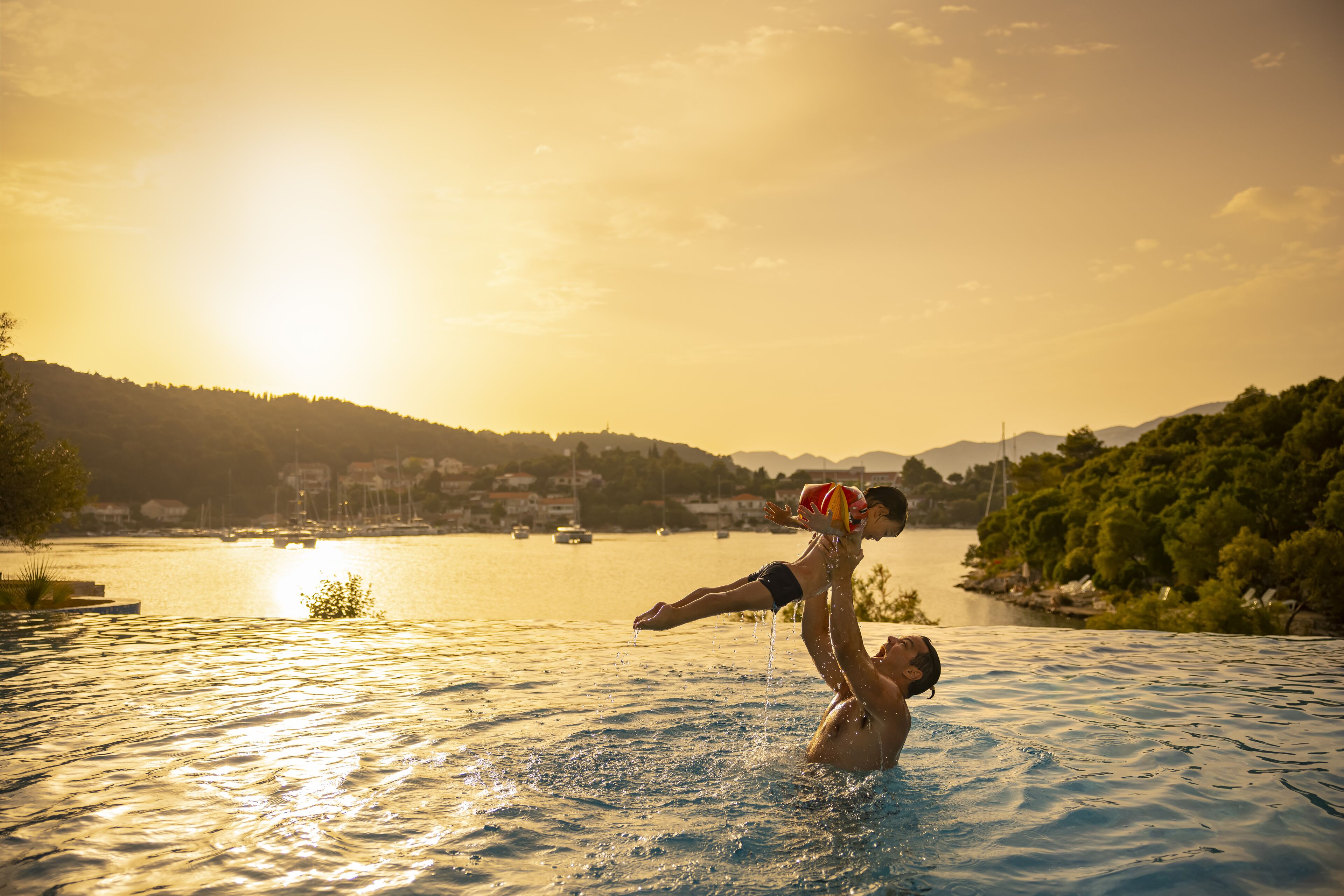 Un hombre levanta a un niño en una piscina con un atardecer de fondo.
