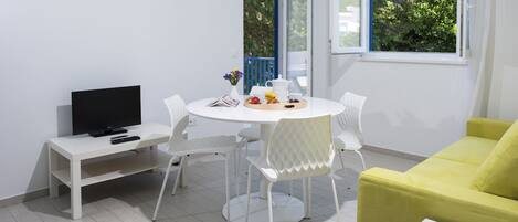 Une pièce moderne avec une table ronde blanche, des chaises blanches, un canapé jaune, un téléviseur posé sur un meuble blanc et une fenêtre donnant sur un paysage verdoyant.