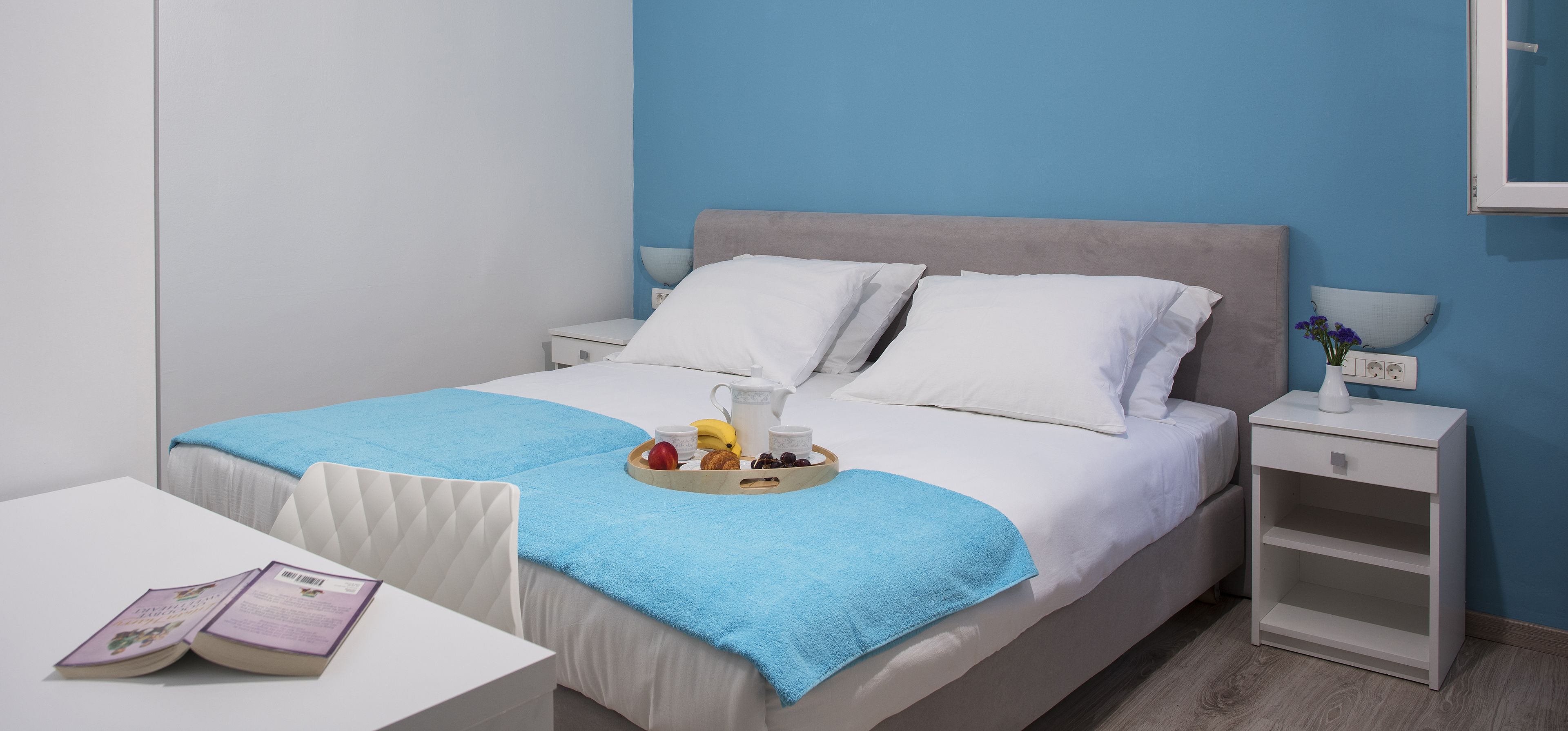 Una camera d'albergo ordinata con un letto, comodini, un tavolino con un vassoio di frutta e un libro, e una parete blu.