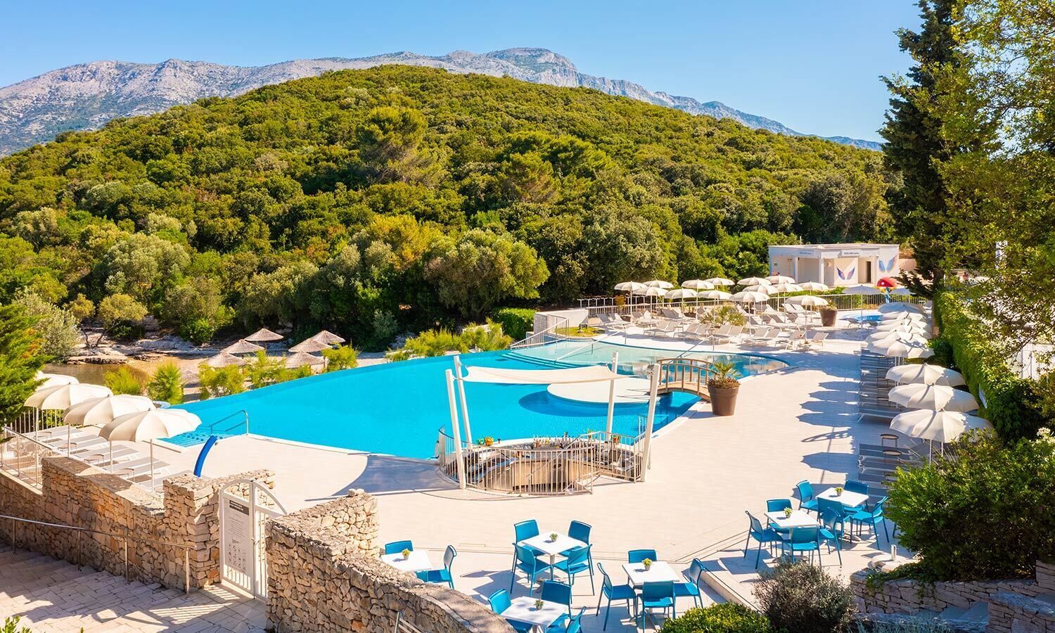 Un espace piscine agrémenté de chaises longues et de parasols, entouré d’un mur de pierre et d’arbres.
