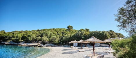A pebble beach with lounge chairs and thatched umbrellas, surrounded by trees and a clear blue sky.