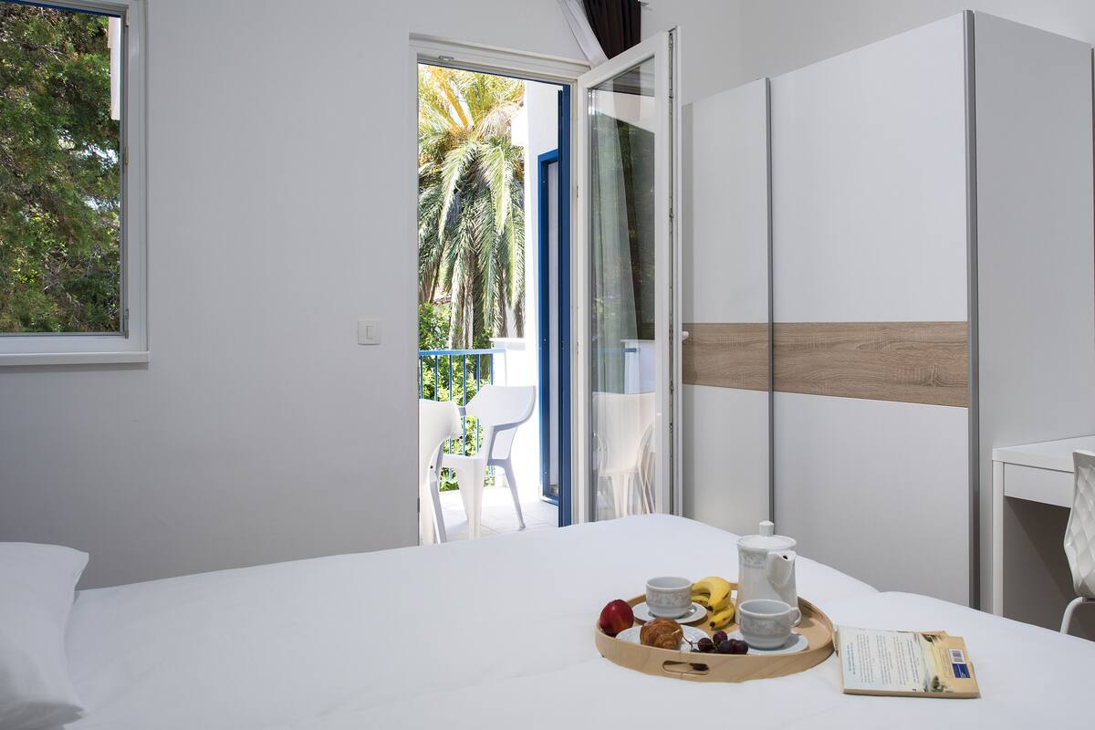 A hotel room with a bed, a dining table with breakfast items, a white chair, and a view of the outside balcony.