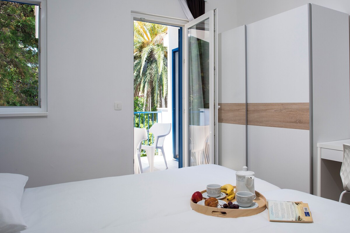 A hotel room with a bed, a dining table with breakfast items, a white chair, and a view of the outside balcony.