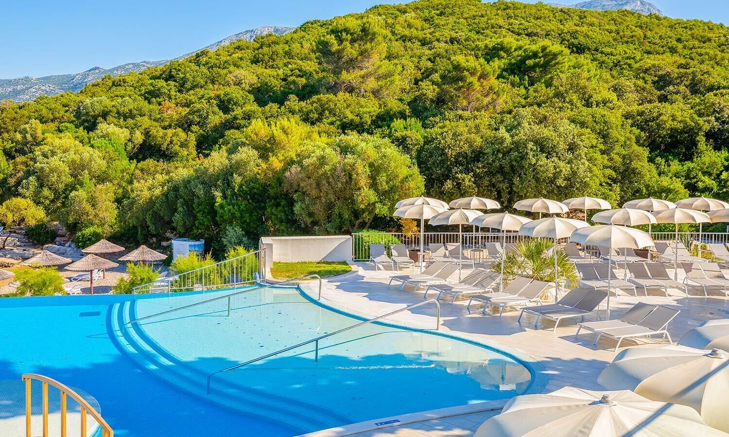 Un espace piscine agrémenté de chaises longues et de parasols, entouré d’une végétation luxuriante et avec une chaîne de montagnes en toile de fond.