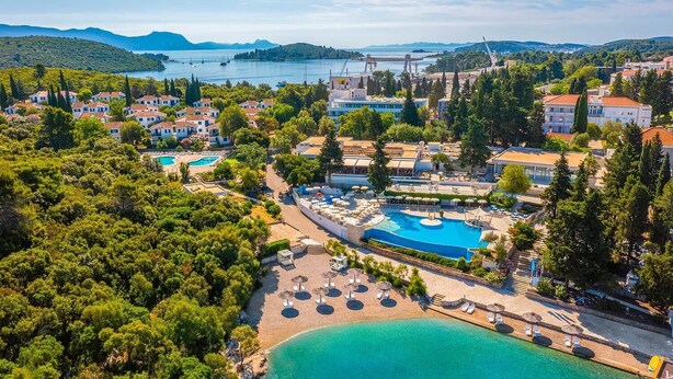 Vista aérea de un complejo turístico costero con varias piscinas, abundante vegetación y colinas con un puente al fondo.