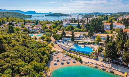 An aerial view of a coastal resort with multiple swimming pools, lush greenery, and a backdrop of hills and a bridge.