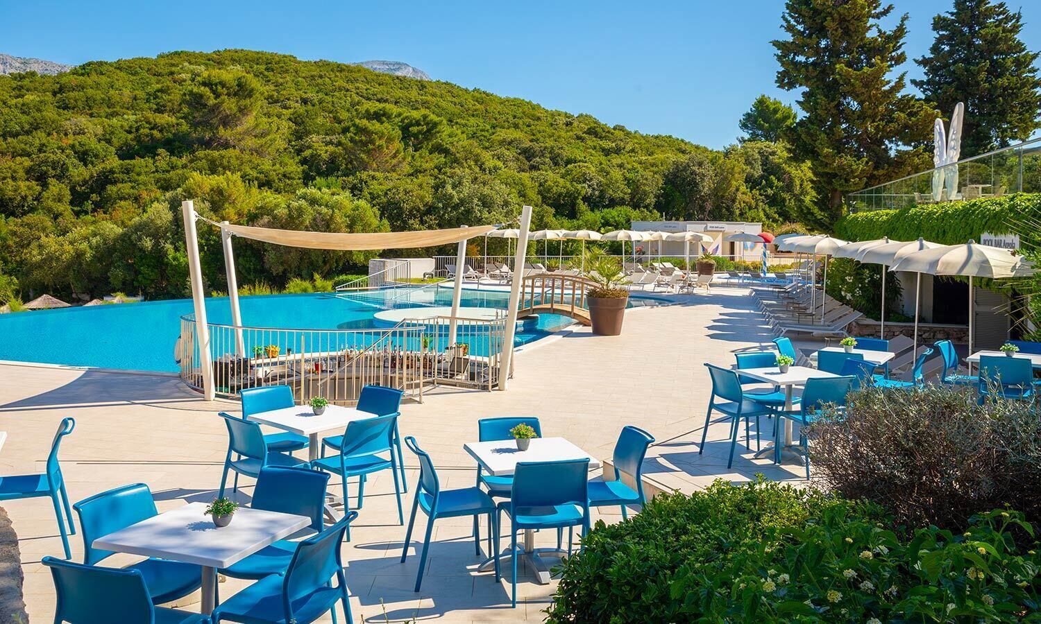 A poolside area with blue chairs and tables, a pool, and a view of green hills.