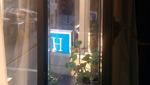 Une vue à travers une fenêtre, avec une enseigne bleue, des plantes en pot et le reflet de bâtiments.