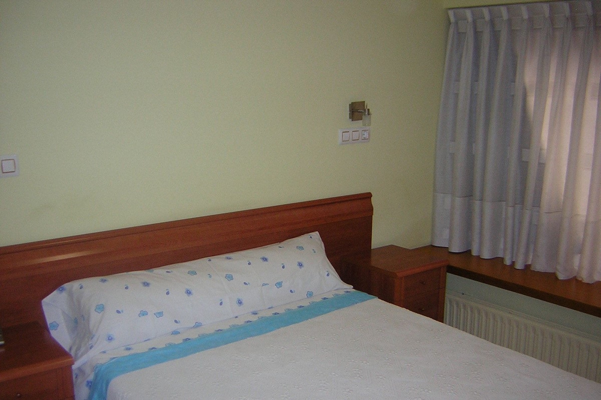 A single bed with a wooden headboard, a white pillow with a blue pattern, and a blue blanket. There is a wooden nightstand to the left and a radiator below the bed.