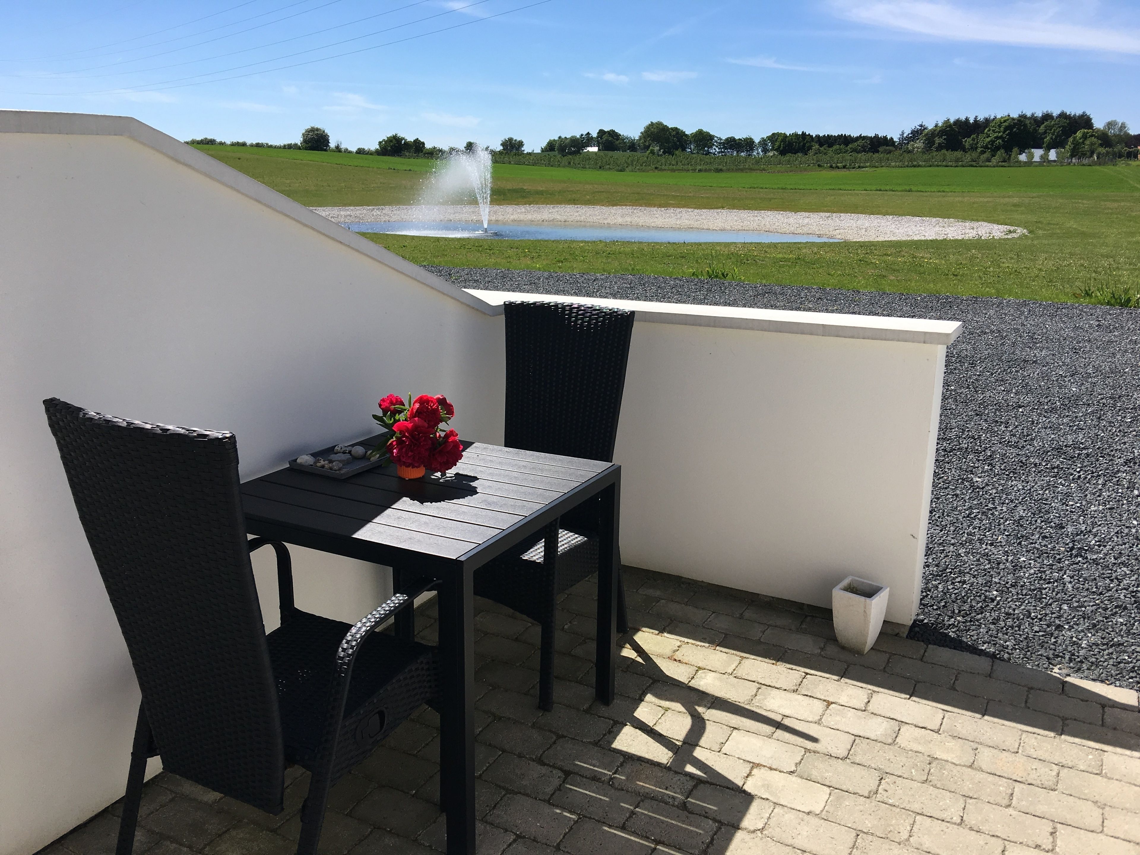 A small outdoor seating area with a table and two chairs, a flower arrangement, and a view of a fountain and green fields.