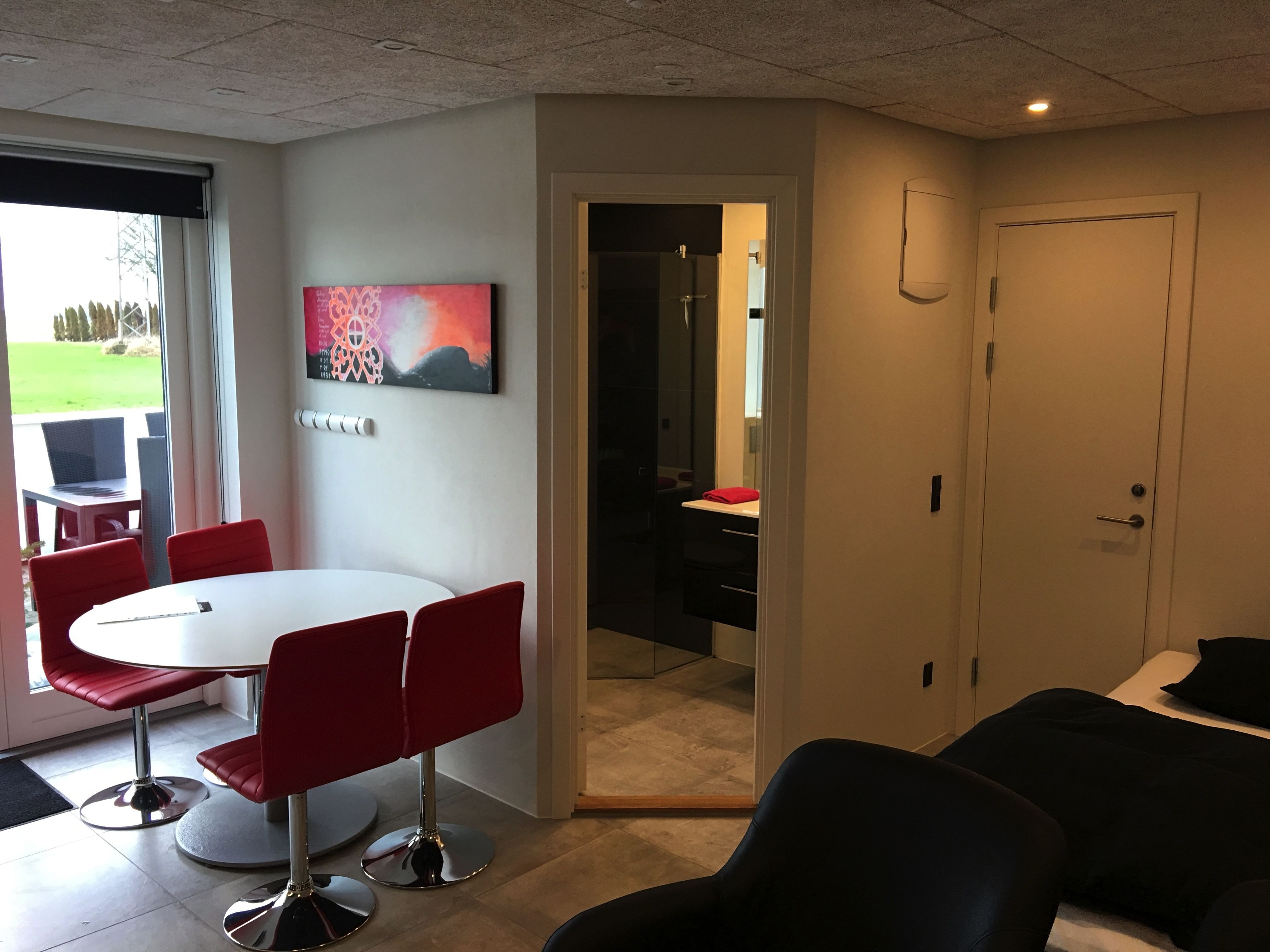 A modern room with a white round table and red chairs, a black sofa, and a door leading to a bathroom.
