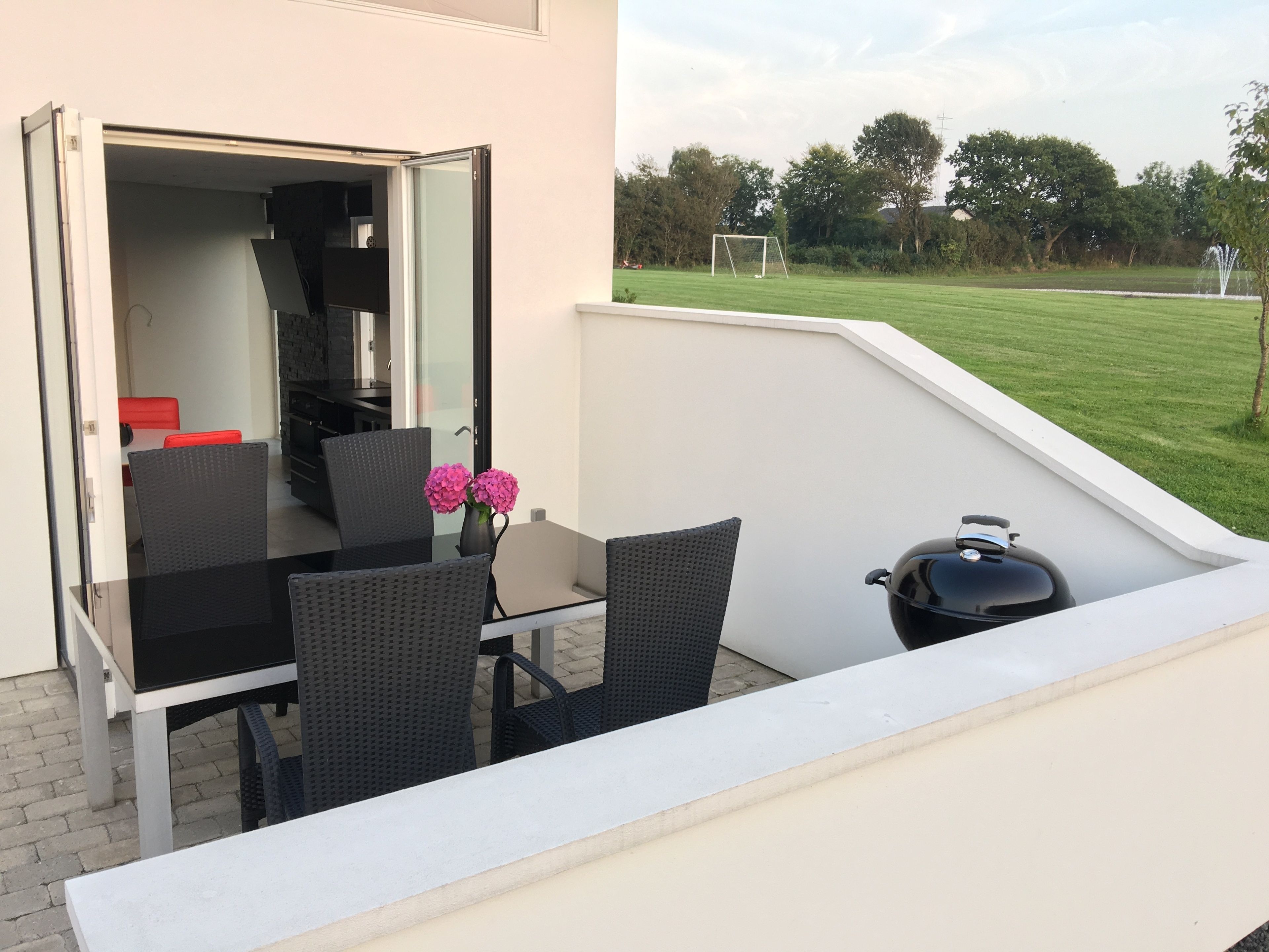 A balcony with a table and chairs, a grill, and a view of a grassy field with a goalpost.