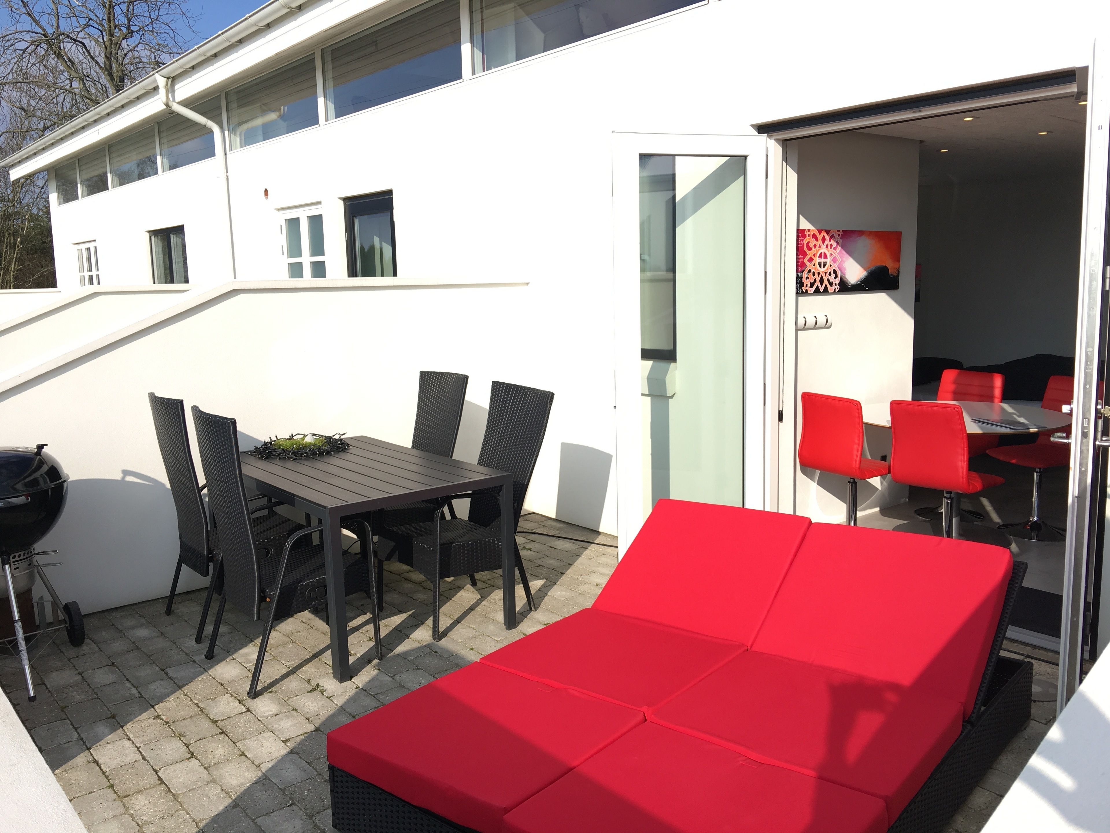 Un patio con una mesa y sillas, una parrilla y una zona de estar con cojines rojos.