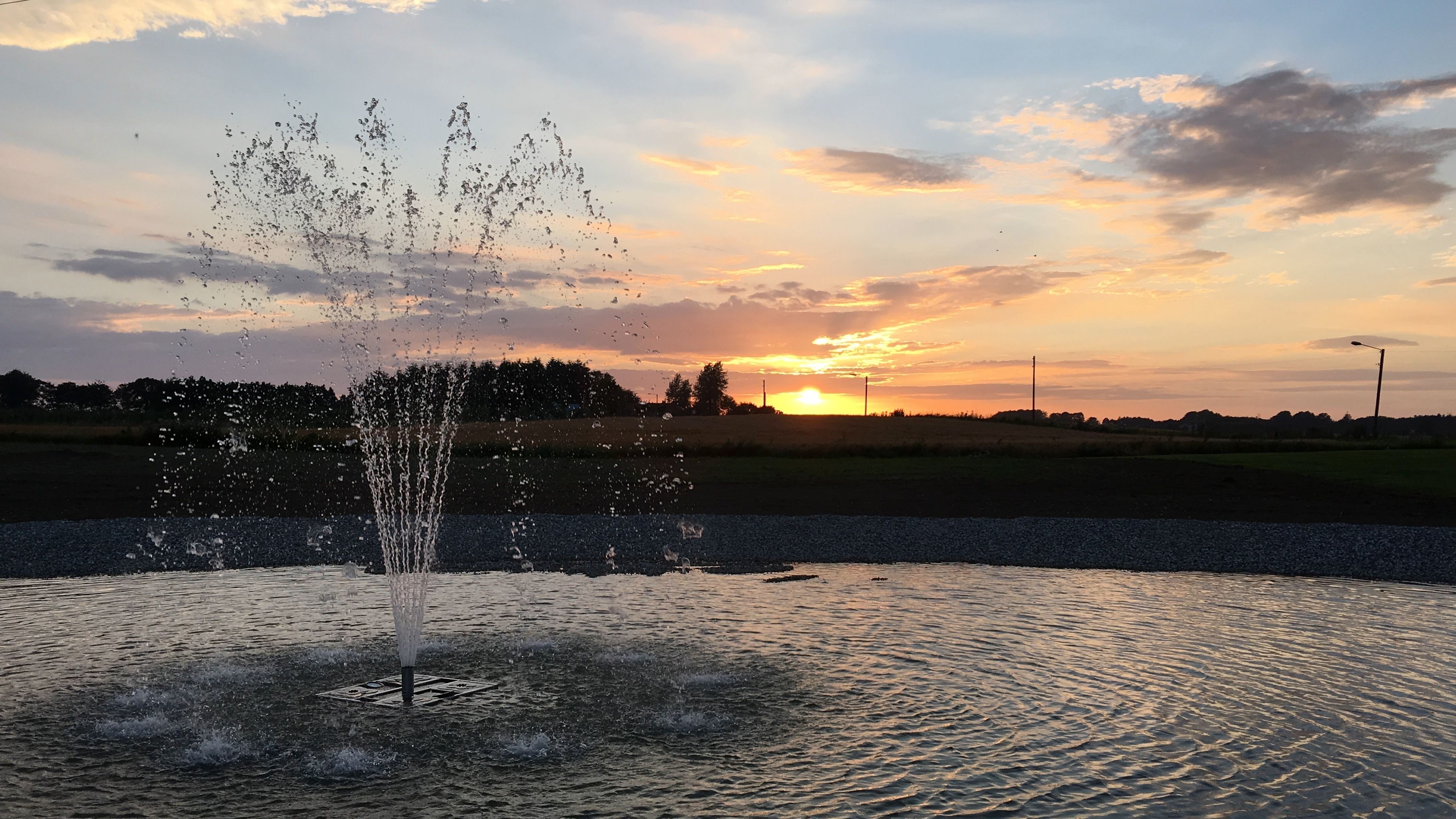 A fountain with water jets in a body of water, set against a sunset sky with clouds.