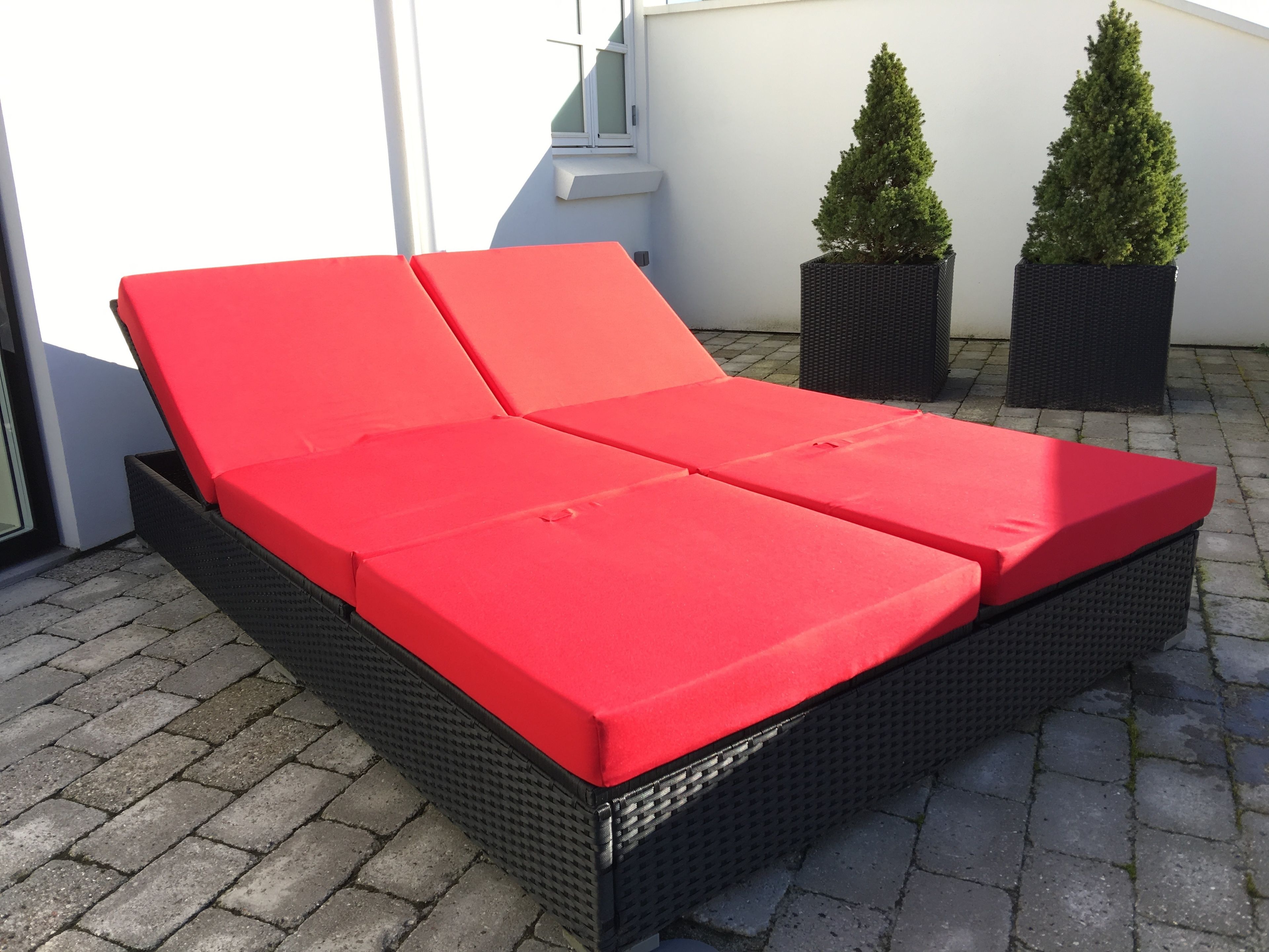A red cushioned outdoor sofa with a wicker frame, placed on a paved patio with two potted evergreen trees in the background.